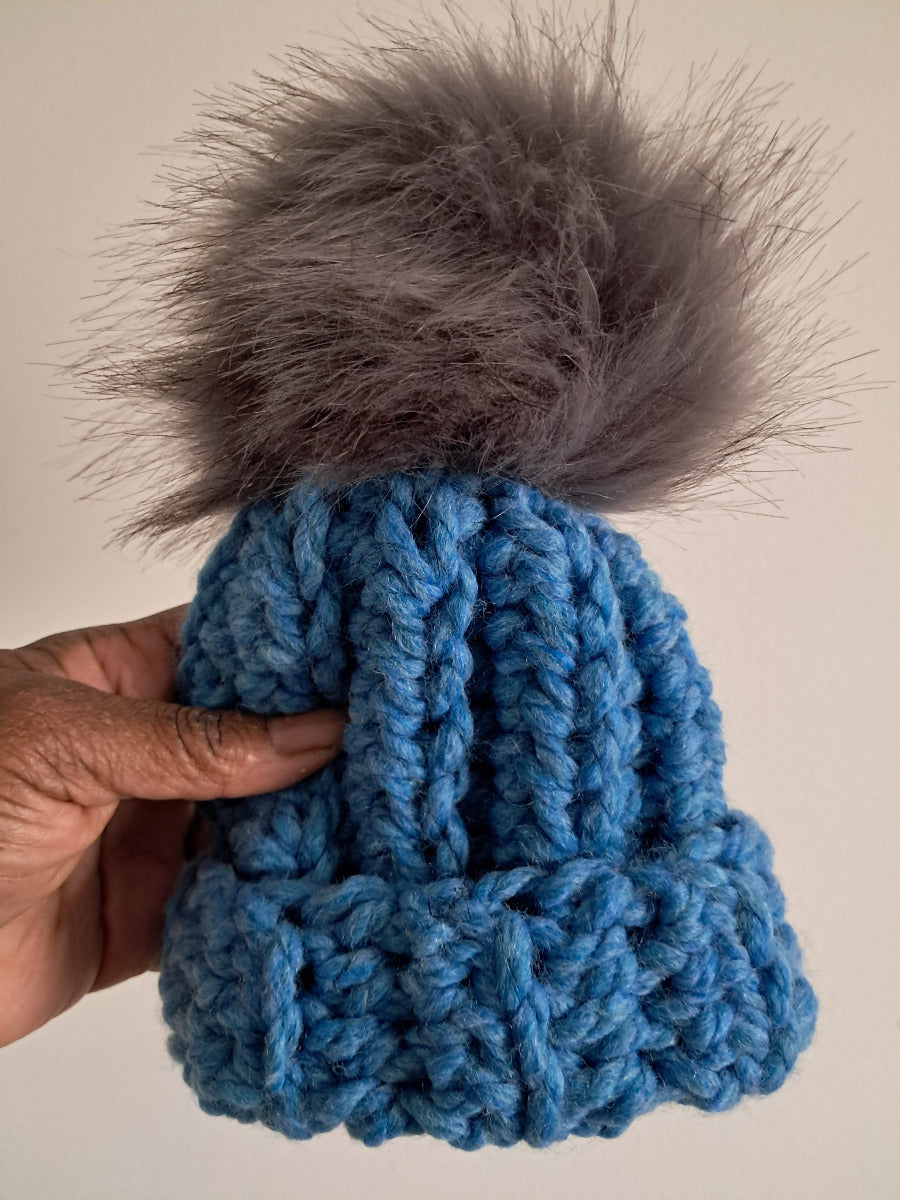 Blue crocheted beanie with a gray pom-pom held by a hand against a plain background