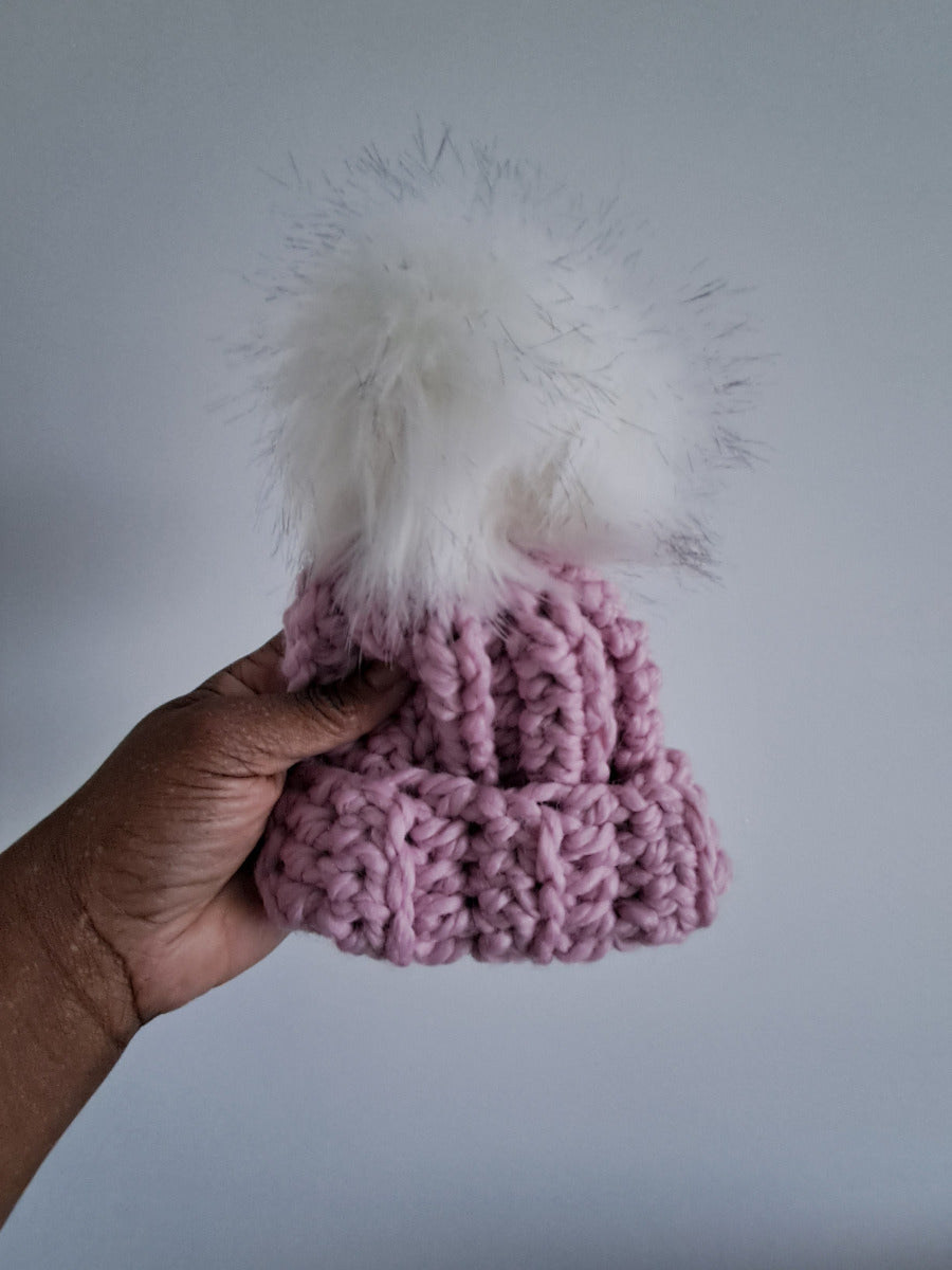 Hand holding a pink crocheted beanie with white fluffy texture pom pom against a plain background