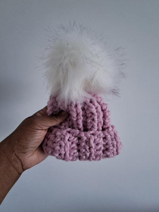 Hand holding a pink crocheted beanie with white fluffy texture pom pom against a plain background