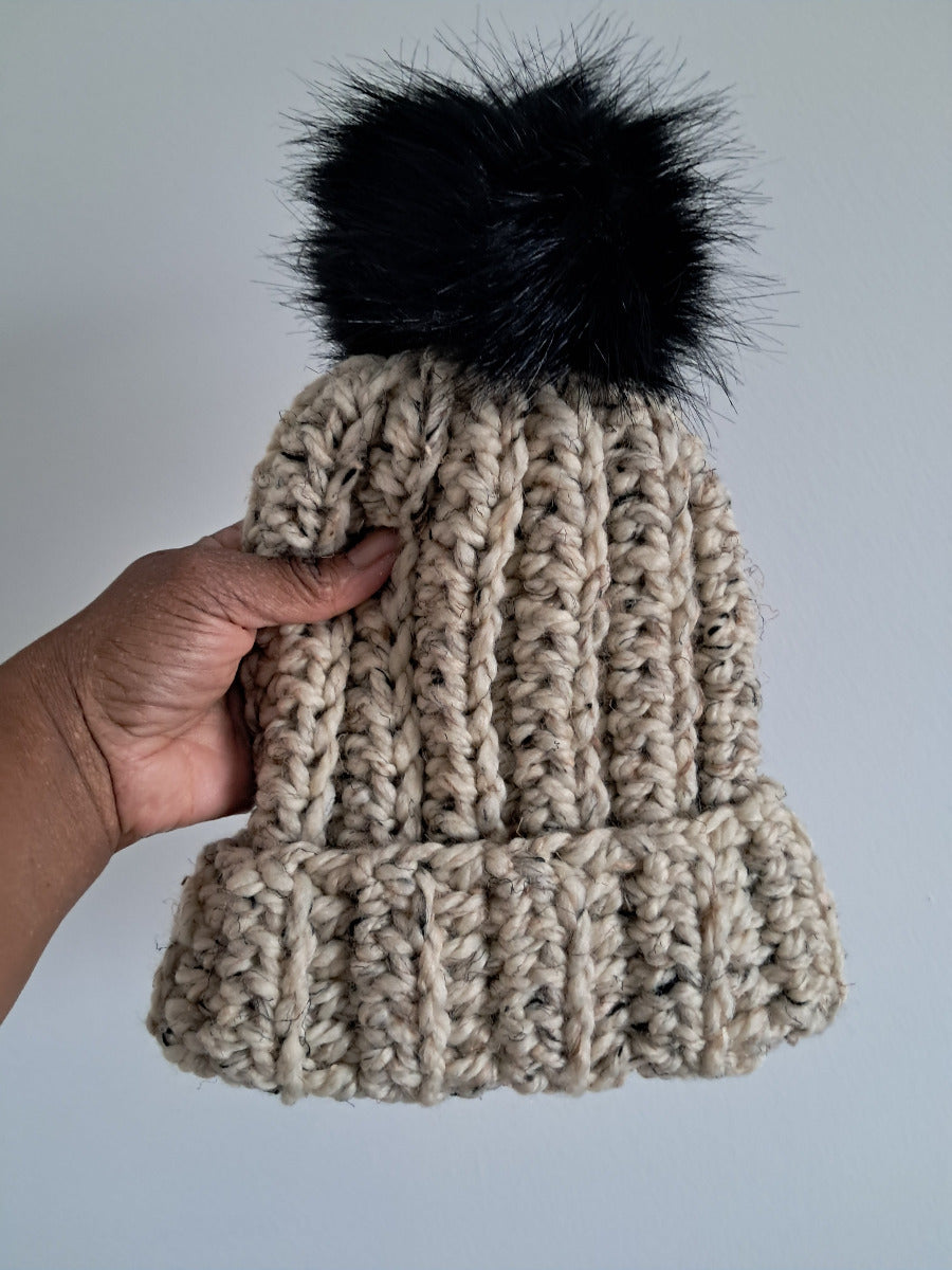 Beige crocheted beanie with a black pom-pom held against a light gray background