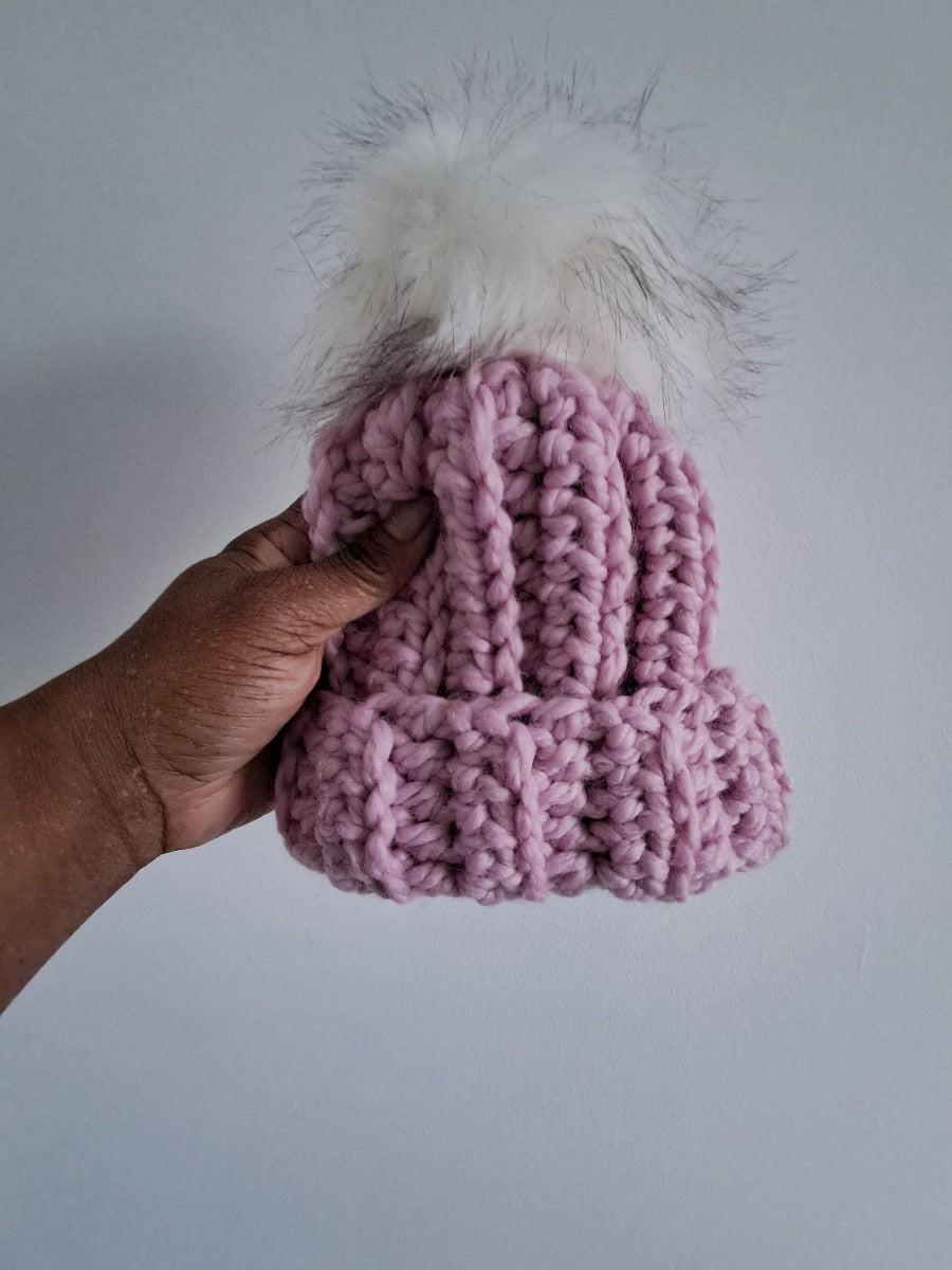 Pink crocheted beanie with a white fluffy pom-pom held by a hand against a light gray background