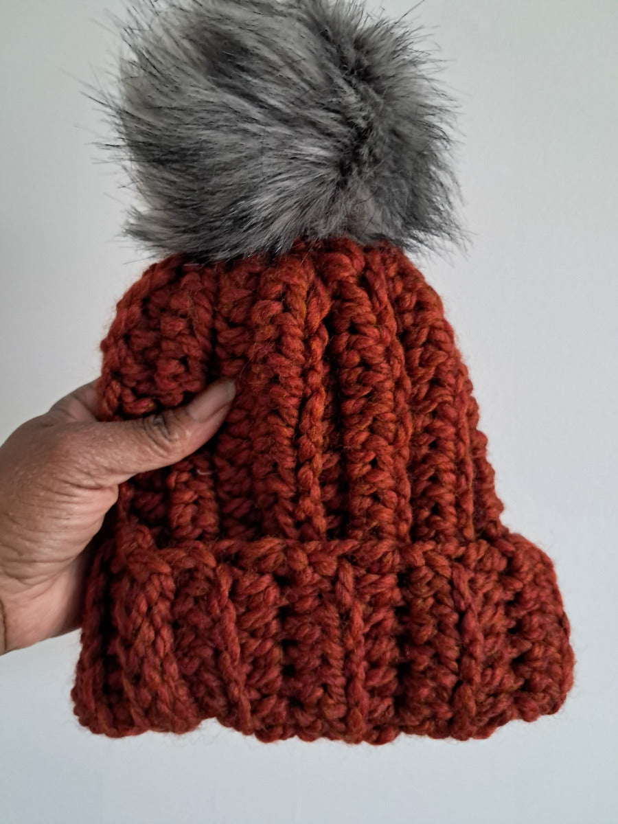 Orange-Red crocheted beanie with a gray pom-pom held by a hand against a light background