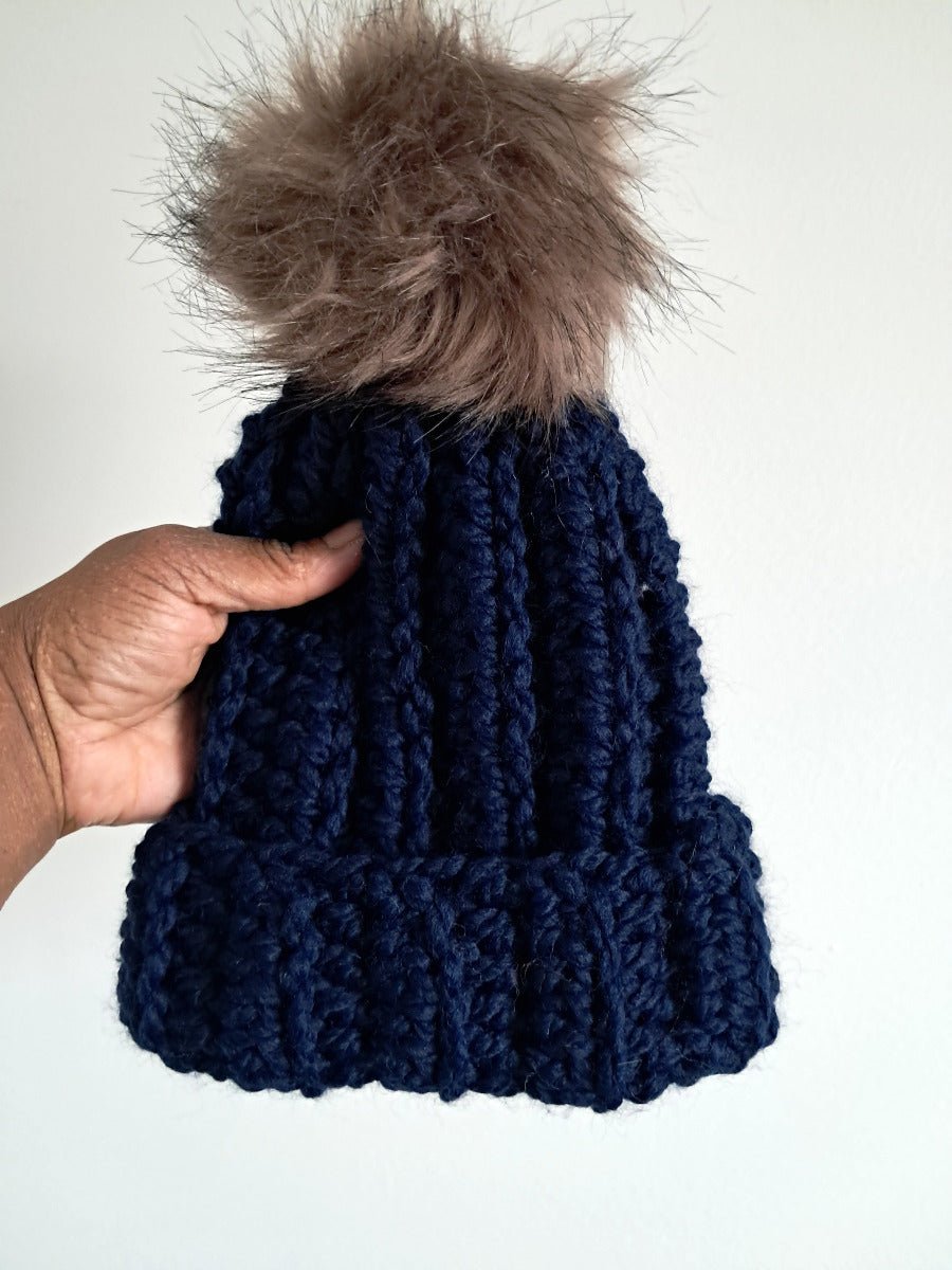 Navy blue crocheted beanie with a fur pom-pom held against a white background