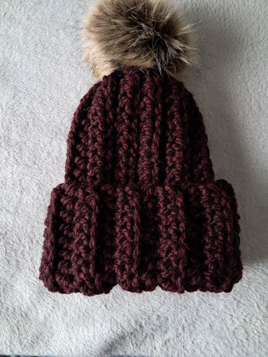 Crocheted maroon beanie with a fur pom-pom on a cream background