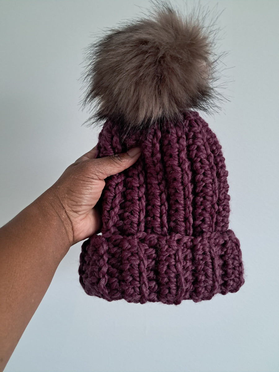 Purple crocheted beanie with a fur pom-pom held by a hand against a light gray background